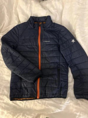 Mörkblå dunjacka från Everest - Snygg mörkblå dunjacka från Everest med orange dragkedja och detaljer. Jackan är quiltad och har hög krage samt två sidofickor med dragkedja. Perfekt för kyliga dagar!