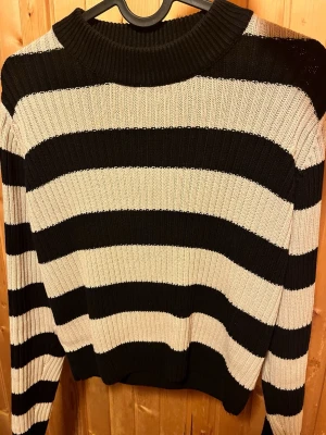 Randig stickad tröja - Stickad tröja från Lager 157 i svart och beige med breda horisontella ränder. Tröjan har ribbstickad struktur, rund halsringning och långa ärmar. Perfekt för dig som gillar klassisk men ändå trendig stil.