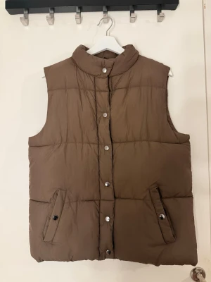 Brun dunväst med tryckknappar - Snygg brun dunväst. Västen har två snedställda fickor med knappstängning och quiltad design. I storlek s, kan bäras av storlek m