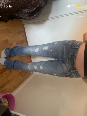 Blå slitna bootcut jeans - Säljer ett par blå bootcut jeans med slitningar och hål på benen. Jeansen har låg midja, klassiska fem fickor och råa kanter vid bensluten. Perfekta för en avslappnad och trendig look.