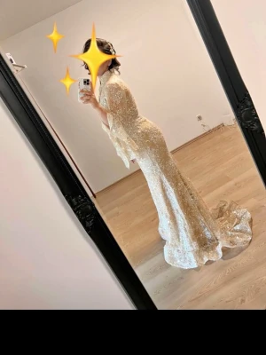 Glittrig långklänning i champagne - Säljer en elegant glittrig långklänning i champagnefärg med lång ärm och utsvängda ärmslut. Klänningen har figurnära passform och vacker släp. Perfekt för speciella tillfällen och bal.