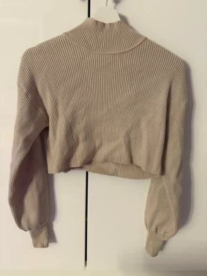 Beige ribbad cropped polo tröja - Trendig beige cropped polo tröja med ribbad struktur och hög krage. Tröjan har långa ärmar med breda muddar och är gjord i ett mjukt material som känns skönt mot huden. Perfekt för dig som gillar en avslappnad men snygg stil.