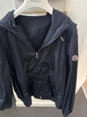 Marinblå vindjacka från Moncler - Snygg marinblå vindjacka från Moncler med huva och dragkedja framtill. Jackan har Moncler-logga på ärmen och diskreta fickor med tryckknappar. Materialet är tunt och lätt, perfekt för blåsiga dagar. Ärmen har en randig detalj i rött, vitt och blått.