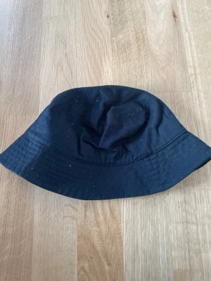 Svart bucket hat i bomull - En stilren svart bucket hat i mjuk bomull med klassisk form och bred brätte. Perfekt accessoar för en avslappnad streetwear-look. Inga synliga loggor eller märken.