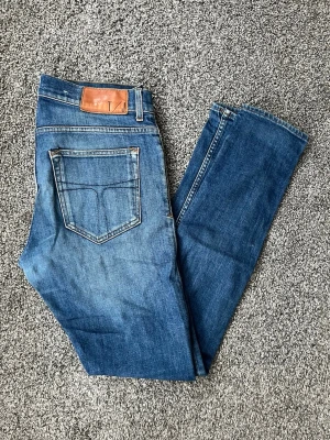 Blå slim fit jeans från Tiger of Sweden - Snygga blå slim fit jeans från Tiger of Sweden med klassisk femficksdesign och läderpatch bak i midjan. Jeansen har en lätt tvättad look och smal passform som sitter tajt längs benen. Perfekta för en trendig och avslappnad stil.