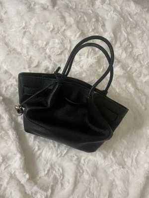 Svart handväska med tillhörande axelrem - Snygg svart handväska i strukturerat skinn med dubbla handtag och avtagbar axelrem. Väskan har en modern, mjuk form och stängs med dragkedja. En dekorativ knutdetalj och silverfärgad berlock hänger på ena sidan( går att ta bort). Perfekt för dig som gillar stilrena accessoarer.
