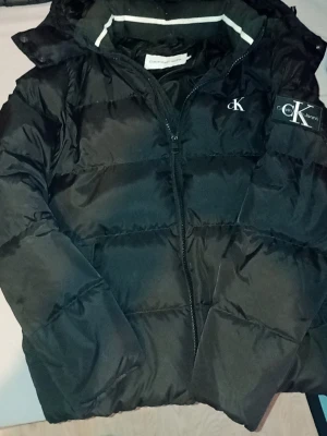 Svart pufferjacka från Calvin Klein - Snygg svart pufferjacka från Calvin Klein med dragkedja framtill och logga på bröstet samt ärmen. Jackan har hög krage och vadderad design som håller dig varm under kalla dagar. Perfekt för vintern och riktigt trendig stil.