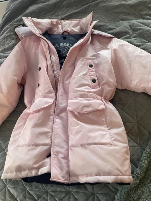 Ljusrosa parkas med fuskpäls - Snygg ljusrosa parkas med dragkedja och tryckknappar framtill. Jackan har stora fickor med lock och en huva med fuskpäls. Perfekt för kyliga dagar och har en lätt figursydd passform. Materialet är glansigt och mjukt. Strl 134 9-10 år 