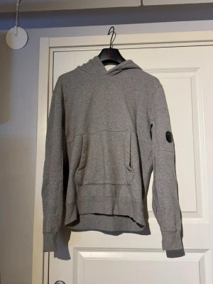 Grå hoodie från C.P. Company - Snygg grå hoodie från C.P. Company med klassisk huva och magficka. Ärmen har det ikoniska linsmärket som ger en unik touch. Jag säljer denna då min pojkvän använt denna ca 3 gånger. Original pris 2200kr men jag säljer den för 1100