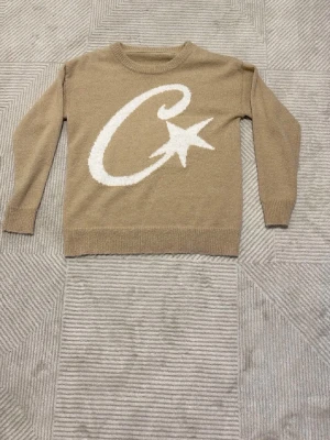 Beige stickad tröja från corteiz - Stickad beige tröja med rund hals och långa ärmar. På framsidan finns ett stort vitt motiv med en snirklig bokstav och en stjärna. Tröjan har ribbade muddar vid ärmslut och nederkant. Perfekt för dig som gillar grafiska detaljer. Priset är diskutabelt