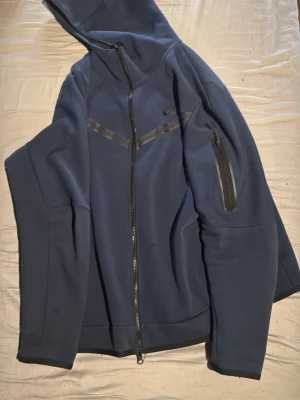 Mörkblå Nike hoodie med dragkedja - Snygg mörkblå hoodie från Nike med hel dragkedja framtill och huva. Har svarta detaljer i form av ett V-format band över bröstet och en praktisk ficka med dragkedja på ena ärmen. Tillverkad i ett mjukt och stretchigt material som känns sportigt.