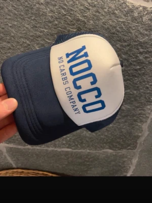 Blå och vit NOCCO keps - Snygg keps från NOCCO med vit frontpanel och blått nät samt skärm. Stor blå logga och texten 'NO CARBS COMPANY' framtill. Klassisk truckerkeps med böjd skärm och justerbar baksida. Perfekt för dig som gillar sportig och avslappnad stil.