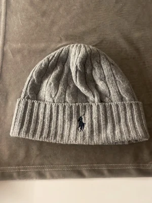 Grå kabelstickad mössa Polo Ralph Lauren - Snygg grå kabelstickad mössa från Polo Ralph Lauren med uppvikt kant och klassisk broderad logga framtill. Tillverkad i mjukt material som håller dig varm och stilsäker under kalla dagar. Perfekt accessoar för en clean och tidlös look.