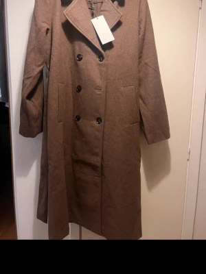 Brun ullkappa från Kaffe - Stilren brun ullkappa från Kaffe med dubbelknäppning och bred krage. Kappan har två stora fickor framtill och en rak, klassisk passform. den är ny med prislapp också 