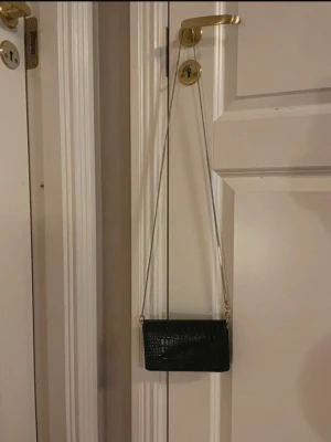 Svart kuvertväska från Gina Tricot - Stilren svart kuvertväska från Gina Tricot med croco-mönstrad yta och lång silverfärgad kedja. Väskan är rektangulär och har magnetlås. Perfekt för att bära det viktigaste när du vill ha en trendig accessoar.