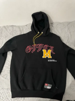 Svart hoodie med tryck från MNML - Svart hoodie från MNML med stor huva och gula dragsnören. Framsidan har ett tryck med röd text 'OFFCL' och ett stort gult M. Känguruficka framtill och ribbade muddar. Materialet känns mjukt och bekvämt, perfekt för en avslappnad streetwear-look.