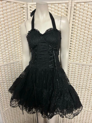 Svart spetsklänning med snörning från Poizen storlek L. - Supersnygg svart kort klänning med spetsdetaljer och snörning framtill. Klänningen har halterneck-band som knyts i nacken och volangig kjol. Perfekt för dig som gillar en edgy och romantisk stil. Helt ny i storlek L. Dragkedja i sidan. Ordpris 899kr.