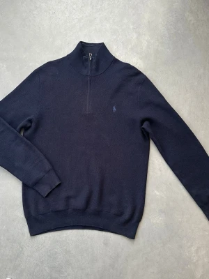 Mörkblå halvzip från Polo Ralph Lauren - Klassisk mörkblå tröja med halv dragkedja från Polo Ralph Lauren. Tröjan har hög krage, ribbade muddar och diskret broderad logga på bröstet. Perfekt för en stilren och avslappnad look. Materialet är mjukt och bekvämt. Köpt några år sedan. Bra skick. 