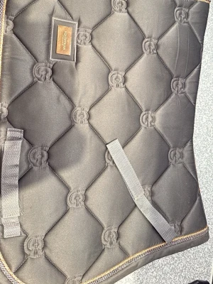 Svart schabrak från Equestrian Stockholm - Snyggt svart schabrak från Equestrian Stockholm med quiltad design och broderad logga. Schabraket har guldfärgade detaljer och praktiska remmar för fastsättning. Perfekt för ridning och ger en stilren look till din häst.