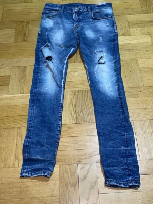 Blå slitna jeans från Dsquared2 - Säljer ett par blå jeans från Dsquared2 med slitningar och hål på båda benen. Jeansen har skinny fit, klassisk femficksmodell och tydliga tvättade detaljer. Röd Dsquared2-logga vid gylfen och kontrastsömmar ger en extra snygg touch.