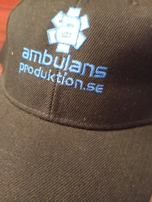 Svart keps Ambulansproduktion.se - Svart keps med böjd skärm och broderad logga i ljusblått och vitt från Ambulansproduktion.se framtill. Klassisk form med ventilationshål upptill och justerbar passform. Materialet är bomull och kepsen har en stilren look som funkar till många outfits.