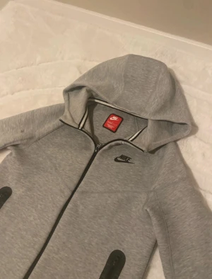 Grå hoodiejacka från Nike - Grå hoodiejacka från Nike med svart logga på bröstet och svarta detaljer vid fickorna. Jackan har dragkedja framtill, huva och långa ärmar. Perfekt för chill eller sport, med mjukt material och stilren look.
