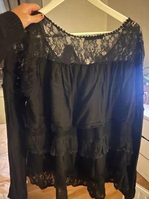 Svart blus med spets och volanger - Säljer en svart blus med lång ärm och snygga spetsdetaljer upptill och längs nederkanten. Blusen har flera lager med volanger som ger en cool och romantisk vibe. Perfekt för dig som gillar att sticka ut med detaljer och älskar svart.