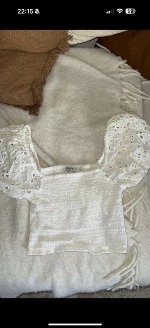 Vit blus med puffärm från NA-KD - Supersöt vit blus från NA-KD med croppad passform och puffiga kortärmar i broderad spets. Toppen har fyrkantig halsringning och smockad struktur i kroppen. Perfekt för dig som gillar romantiska detaljer och vill sticka ut lite extra.