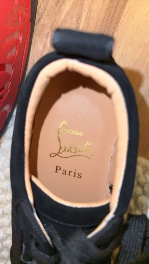 Christian Louboutin svarta sneakers - Svarta sneakers från Christian Louboutin med ikonisk röd sula och snygg signatur. Skorna har rund tå, platt sula och snörning. Ovandelen är i mjuk mocka och insidan är beige med guldfärgad logga. Perfekt för dig som gillar lyxiga detaljer och stilren design.