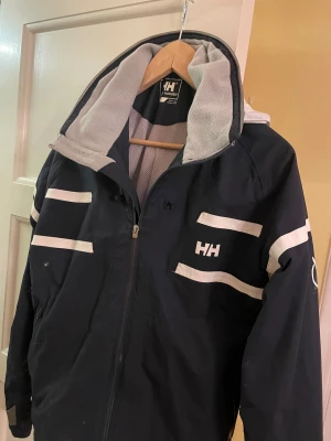 Svart vindjacka från Helly Hansen  - Svart vindjacka från Helly Hansen med vita ränder och HH-logga på bröstet. Jackan har dragkedja framtill, hög krage med grått fleecefoder och sportig design. Perfekt för dig som gillar funktionella och snygga ytterplagg med coola detaljer. För mer frågor skriv privat!