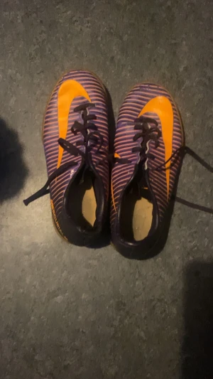 Nike fotbollsskor lila och orange - Säljer ett par Nike fotbollsskor med lila och orange randigt mönster och stor orange swoosh på sidan. Skorna har snörning och platt sula i gummi, perfekt för inomhusspel. Snygg design som sticker ut på planen.
