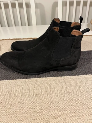 Svarta chelsea boots från H&M, stl 43 - Snygga svarta chelsea boots från H&M i storlek 43. Skorna har en klassisk siluett med elastiska paneler på sidorna och dragflik bak. Ovandelen är i mockaliknande material och sulan är platt med låg klack. Perfekta för en stilren look. Använd bara 1 gång.