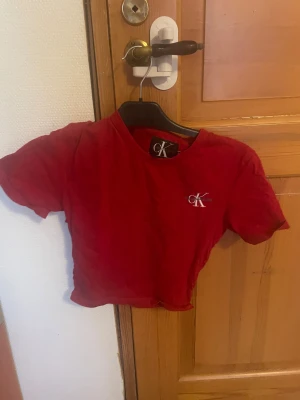 Röd croppad topp från Calvin Klein - Säljer en röd croppad topp från Calvin Klein Jeans den är sydd så med broderad CK-logga i vitt på bröstet. den är tightare i midjan. T-shirten har rund hals, korta ärmar och är gjord i mjuk bomull. Perfekt för dig som gillar klassisk streetstyle med en modern twist.