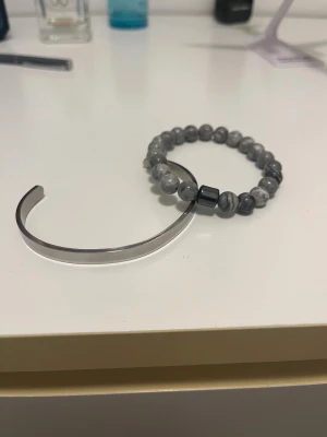 Grått pärlarmband och silverfärgat cuff - Säljer ett stilrent set med ett grått pärlarmband med marmorerade kulor och ett minimalistiskt silverfärgat cuff-armband. Perfekt för dig som gillar att mixa olika stilar och material. Armbanden är enkla att matcha med andra accessoarer. Silver armbandet är justerbart.