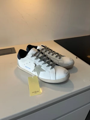 Golden Goose vita sneakers med stjärna - Har tagit av lappen på Golden Goose sneakers i vitt skinn med grå mockadetaljer och grå snörning. Sidan pryds av en ikonisk stjärna i ljus mocka och hälen har en mörkblå läderpatch. Klassisk låg modell med platt sula och diskret branding på sidan.
