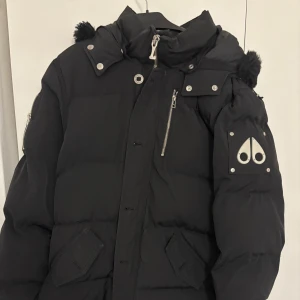 Svart pufferjacka från Moose Knuckles - Svart pufferjacka från Moose Knuckles med ikonisk logga i metall på ärmen, huva med pälsdetaljer och flera dragkedjor. Jackan har långa ärmar, praktiska fickor och coola metalldetaljer. Perfekt för kalla dagar och riktigt snygg streetstil. storleken är XL men känns som L. OBS ÄKTA Vara