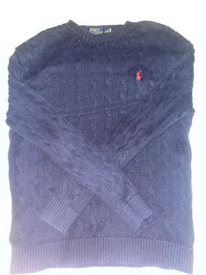 Marinblå stickad tröja Polo Ralph Lauren - Snygg marinblå stickad tröja från Polo Ralph Lauren med klassiskt kabelstickat mönster och röd broderad logga på bröstet. Rund hals och ribbade muddar vid ärmslut och nederkant. Perfekt för dig som gillar stilrena och tidlösa plagg.
