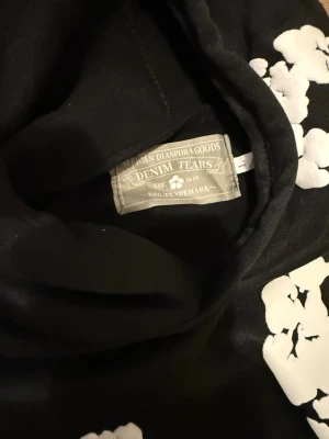Svart hoodie med vita blommor Denim Tears - Svart hoodie från Denim Tears med stora vita blommor tryckta över hela tröjan. Tröjan har huva, magficka och långa ärmar. Materialet är mjukt och känns som bomull, perfekt för dig som vill sticka ut med en unik design.