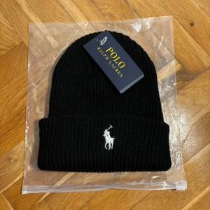 Svart ribbad mössa Polo Ralph Lauren - Svart ribbstickad mössa från Polo Ralph Lauren med klassisk broderad logga i vitt framtill. Tjockt uppvikt kant och stilren design som passar perfekt till kalla dagar. Materialet är mjukt och värmande ull.