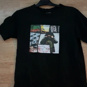 Svart t-shirt Welcome To Sweden XS éinar och 1.cuz - Svart t-shirt från Welcome To Sweden i storlek XS med coolt collage-tryck på bröstet. Trycket har motiv som bil, pengar och maskerad person i olika färger. Klassisk rund hals och korta ärmar. Perfekt för dig som gillar streetwear och grafiska prints. Passar perfekt för dig som har S sitter väldigt bra