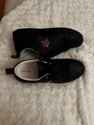 Svarta sneakers från Pier One - Snygga svarta sneakers från Pier One med rund tå och platt sula. Skorna har snörning och ett broderat emblem i guld, rött och blått på sidan samt en diskret mönstrad yta. Insidan är vit och de har en röd-blå detalj på hälen. Perfekt för dig som gillar stilrena detaljer.