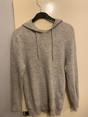 Grå Kashmir hoodie med huva - Mysig grå stickad hoodie med huva och snörning. Tröjan har långärmade ärmar och ribbade muddar vid ärmslut och nederkant. Perfekt för dig som gillar en chill och enkel stil. Passar dig som vill ha något bekvämt och snyggt till vardagen.