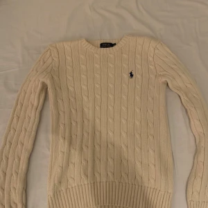 Beige stickad tröja Polo Ralph Lauren - Säljer en beige stickad tröja från Polo Ralph Lauren i storlek XS. Tröjan har klassiskt kabelstickat mönster, rund hals och långärmad design. Den ikoniska Polo-loggan är broderad i mörkblått på bröstet. Perfekt för dig som gillar snygga kläder 
