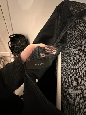 Svart långärmad tröja från Diesel - Svart långärmad tröja från Diesel med cool meshdetalj över axlar och övre rygg. Tröjan har rund halsringning, knappar bak i nacken och broderad Diesel-logga på ärmen. 