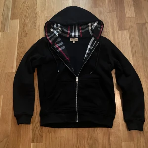 Svart Burberry hoodie  - Svart hoodie från Burberry i 100% bomull med dragkedja framtill och klassisk rutig huva i svart, vitt och rött. Snygg och stilren med snörning i huvan och två fickor framtill. Perfekt för dig som vill ha en lyxig streetstyle-vibe