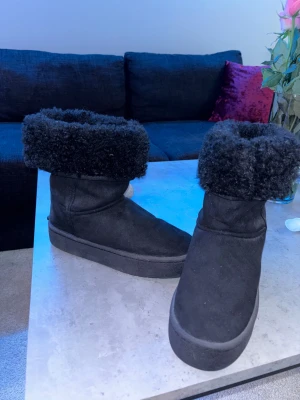 Svarta boots med fluffig kant - Säljer ett par svarta boots med platt sula och rund tå. De har en bred, fluffig kant upptill som ger extra mysig känsla. Utsidan är i mocka och insidan ser ut att vara fodrad för värme. Aldrig använt!