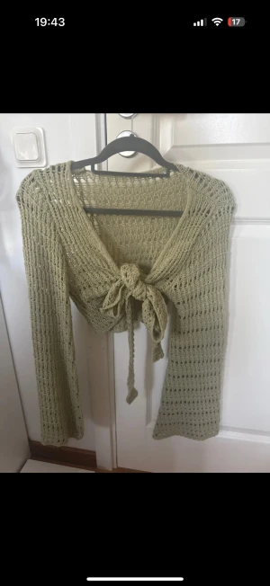 Beige stickad bolerotopp med knyt - Säljer en  stickad bolerotopp med långärmad design och snyggt knyt framtill. Toppen har ett luftigt, hålmönstrat stickat material som ger en chill och trendig vibe. Perfekt att slänga över en linne eller klänning för extra stilpoäng.