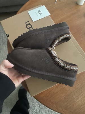 Bruna UGG slip-on mockaskor - Mysiga slip-on skor från UGG i mörkbrun mocka med fluffigt foder och dekorativt mönster runt öppningen. Sulan är platt och grovmönstrad för extra grepp. Perfekt för chill dagar och håller fötterna varma med sin mjuka insida.