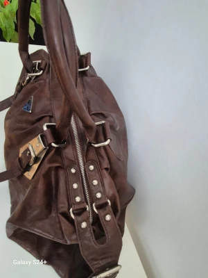 Brun handväska  - Snygg brun handväska  med silverfärgade metalldetaljer och nitar. Väskan har dubbla handtag, dragkedja upptill och coola spännen på framsidan. Perfekt storlek för allt du behöver och riktigt stilren look.