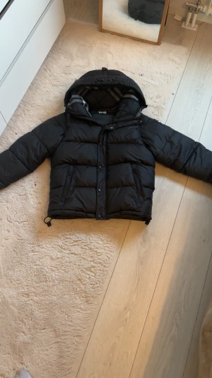  Burberry jacka - Svart pufferjacka från Burberry med huva och klassiskt rutigt foder. Jackan har dragkedja och knappar framtill, två sidofickor med dragkedja och en patch med Burberry London England på ärmen. Perfekt för kalla dagar med sin tjocka vaddering. Helt ny!